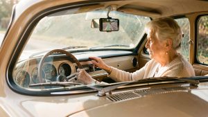 Une femme âgée utilise un smartphone fixé au pare-brise pour la navigation dans une ancienne voiture, sans modifier le tableau de bord.